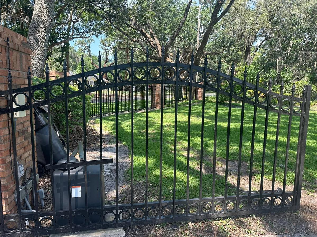 Black arched steel driveway gate with ring detailing, closed across a tree-lined drive